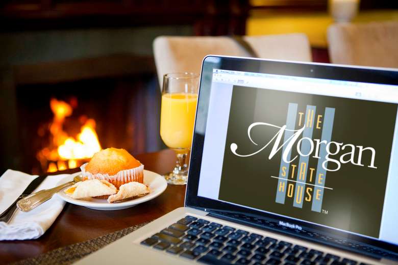 laptop computer with the morgan state house's website pulled up on a table with a pastry and orange juice in front of a fireplace